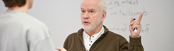 Image of a male professor in a classroom speaking to a student and pointing upward. The back of a student's head is partially in the image.